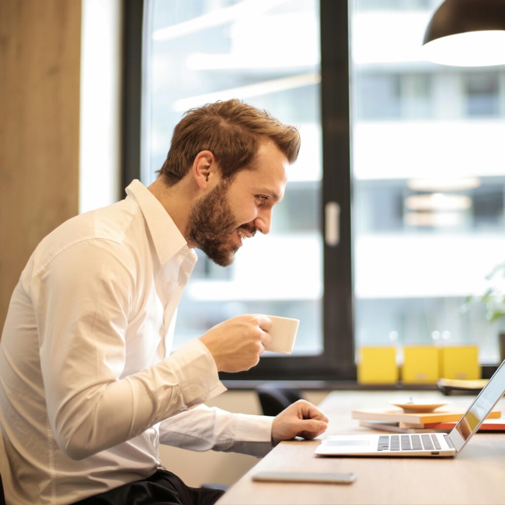 Man in office with coffee, smiling while working at laptop, captures the essence of remote work.